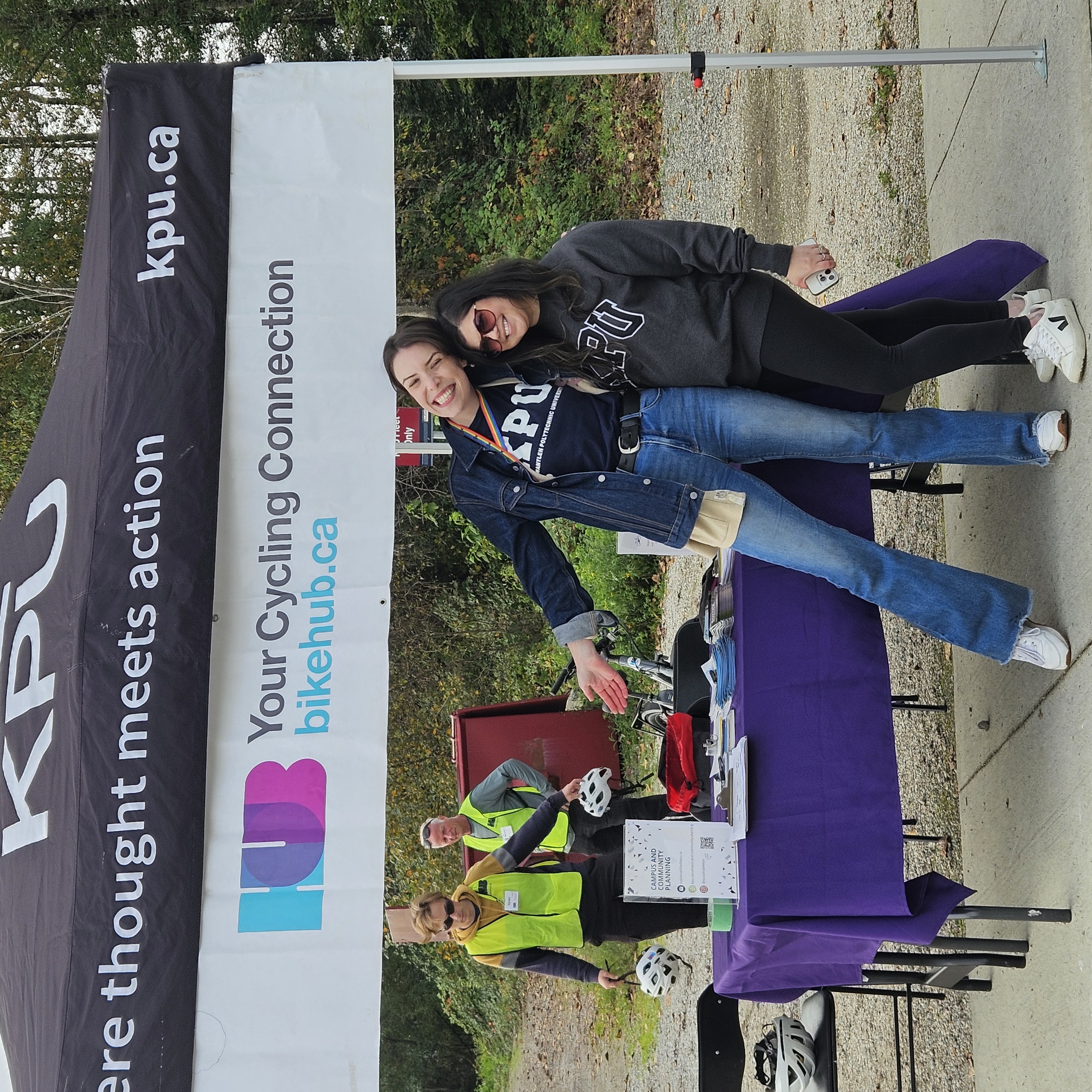 two women wearing KPU sweaters standing infrount of a tent with the KPU logo and Hub Cycling logo