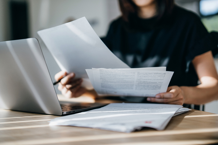 A woman reviewing paper documents sitting at a desk with a laptop.