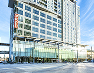 Image of KPU's Civic Plaza Campus, tall and large glass building with blue sky and concrete ground. 