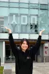 Artist Winnie Hui raises her arms, creating a Y-shape framing the word you in large letters on the first floor of the glass building behind her